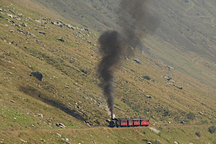 HG4/4 Lok 704 klettert auf der Zahnstange mit dem ersten Zug von Realp nach Oberwald weit unterhalb von Bahnhof Furka den Berg hoch, hier fotografiert im oberen Furkareuss-Tal noch zwischen dem Steintafelsviadukt und der Brücke über den Sidelenbach, um 10:59h am 13.08.2026